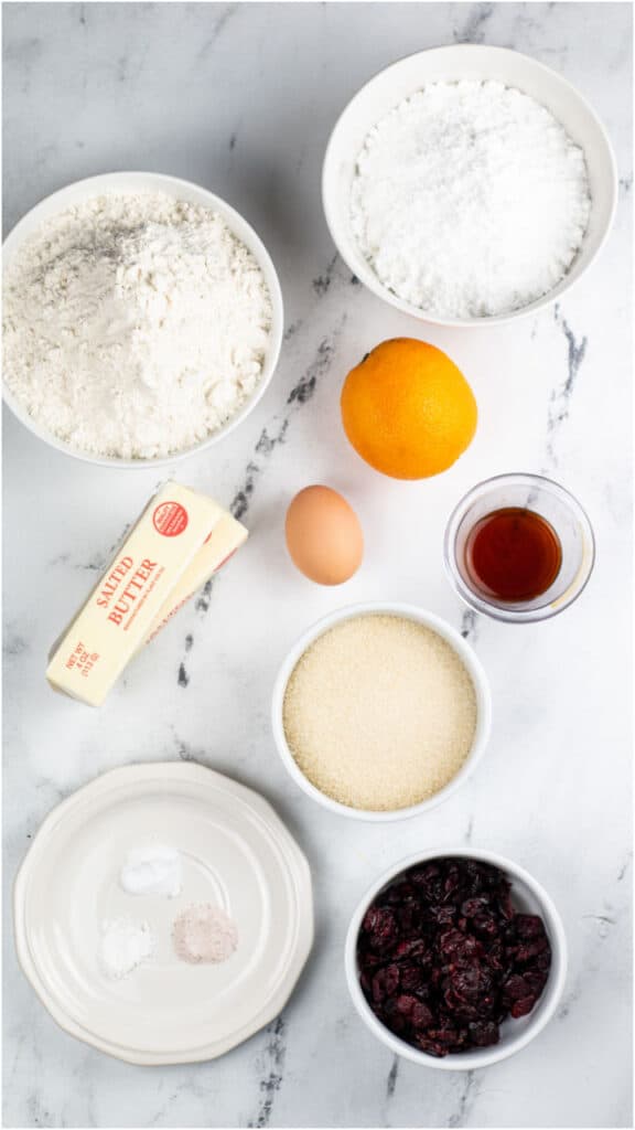The ingredients for cranberry orange cookies on a white marble background. Flour, sugar, orange, egg, vanilla, butter, powdered sugar. dried cranberries, baking soda, baking powder and salt. 