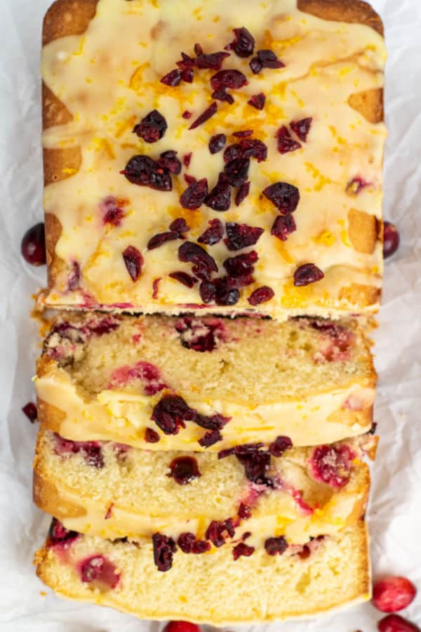 An overhead shot of a cranberry orange pound cake that is glazed with orange glaze and sprinkled with dried cranberries. Half of the cake has been sliced. 