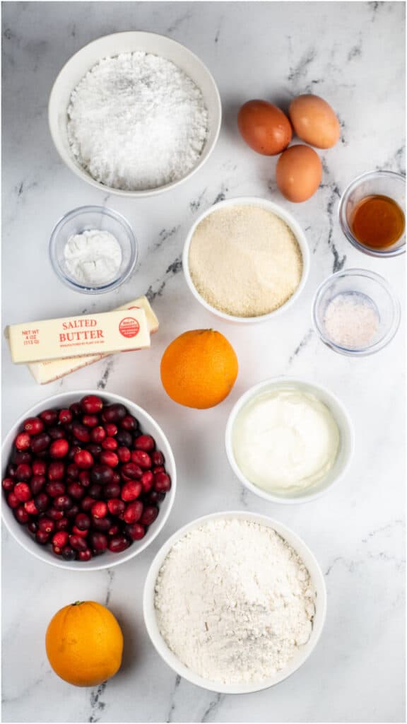 The ingredients for cranberry orange pound cake on a marble countertop. Flour, sugar, eggs, butter, cranberries, sour cream oranges, salt, baking soda, vanilla and powdered sugar. 
