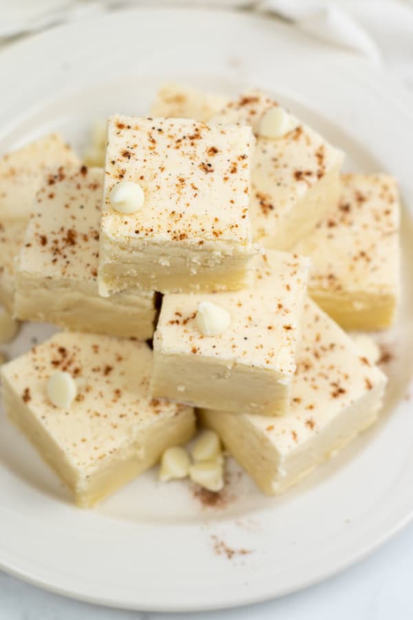 A white plate with a pile of eggnog fudge. The fudge is white and sprinkled with nutmeg. 
