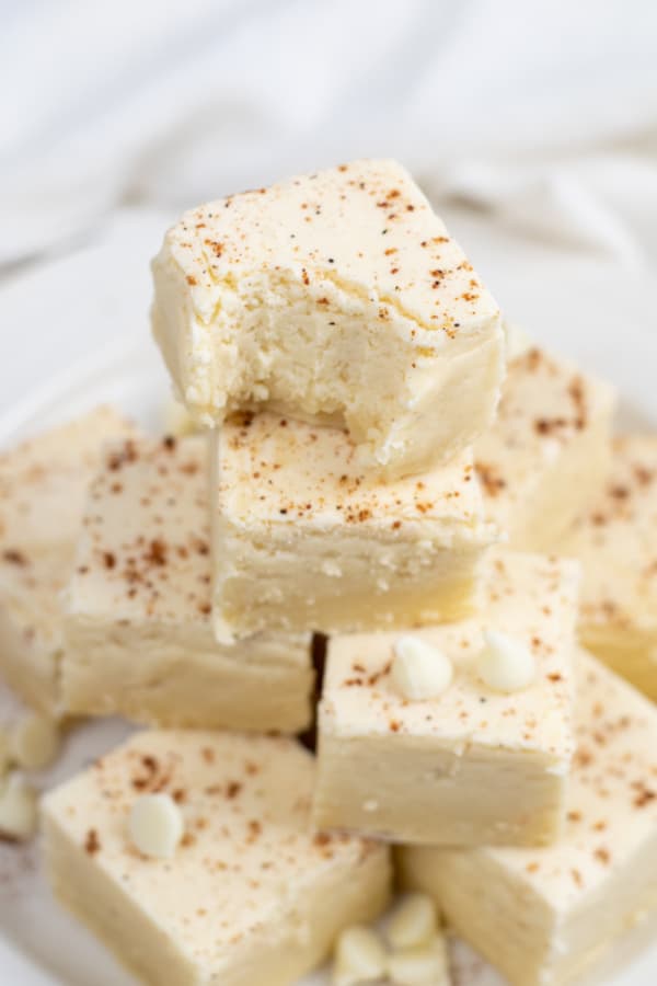 A pile of eggnog fudge on a white plate. The top piece has a bite taken out showing the soft creamy texture of the fudge. 