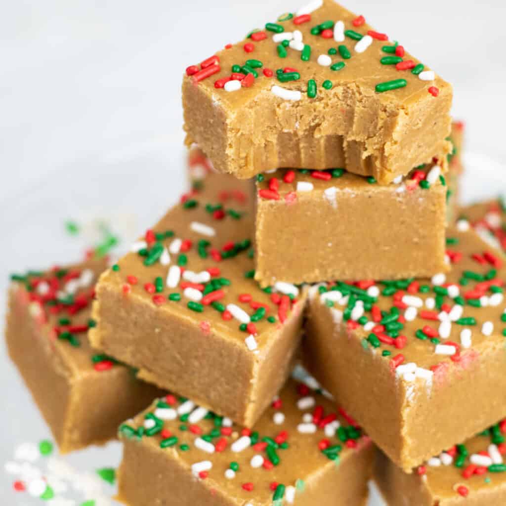 A pile of gingerbread fudge with a bite taken out of the top piece. The fudge is the color of gingerbread and topped with red, green and white sprinkles. 