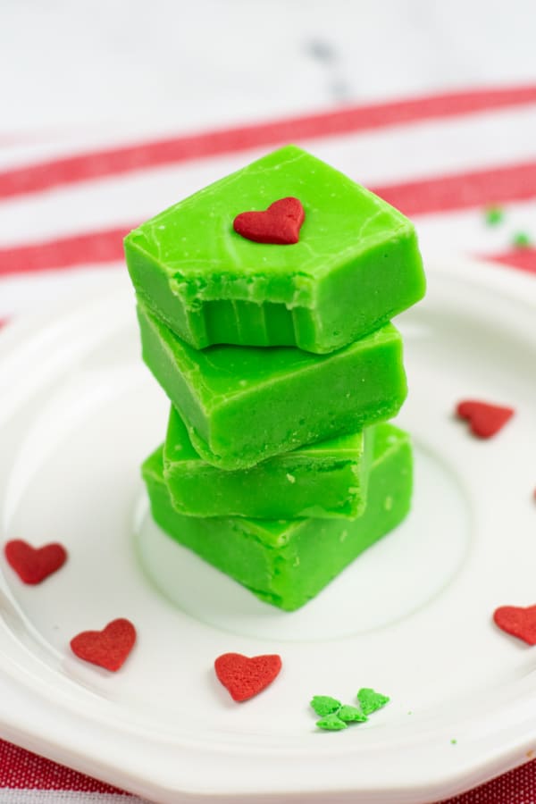 A stack of Grinch fudge on a white plate. The top piece has a bite taken out showing the creamy interior of the fudge. 