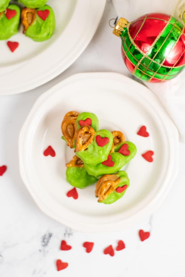 a pile of Grinch pretzel bites on a white plate surrounded by heart shaped sprinkles. 