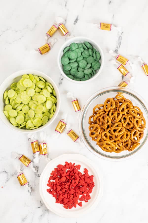 The ingredients for Grinch pretzel bites on a white marble background. Green candy melts, lime green candy melts. pretzels, caramels and heart shaped sprinkles. 