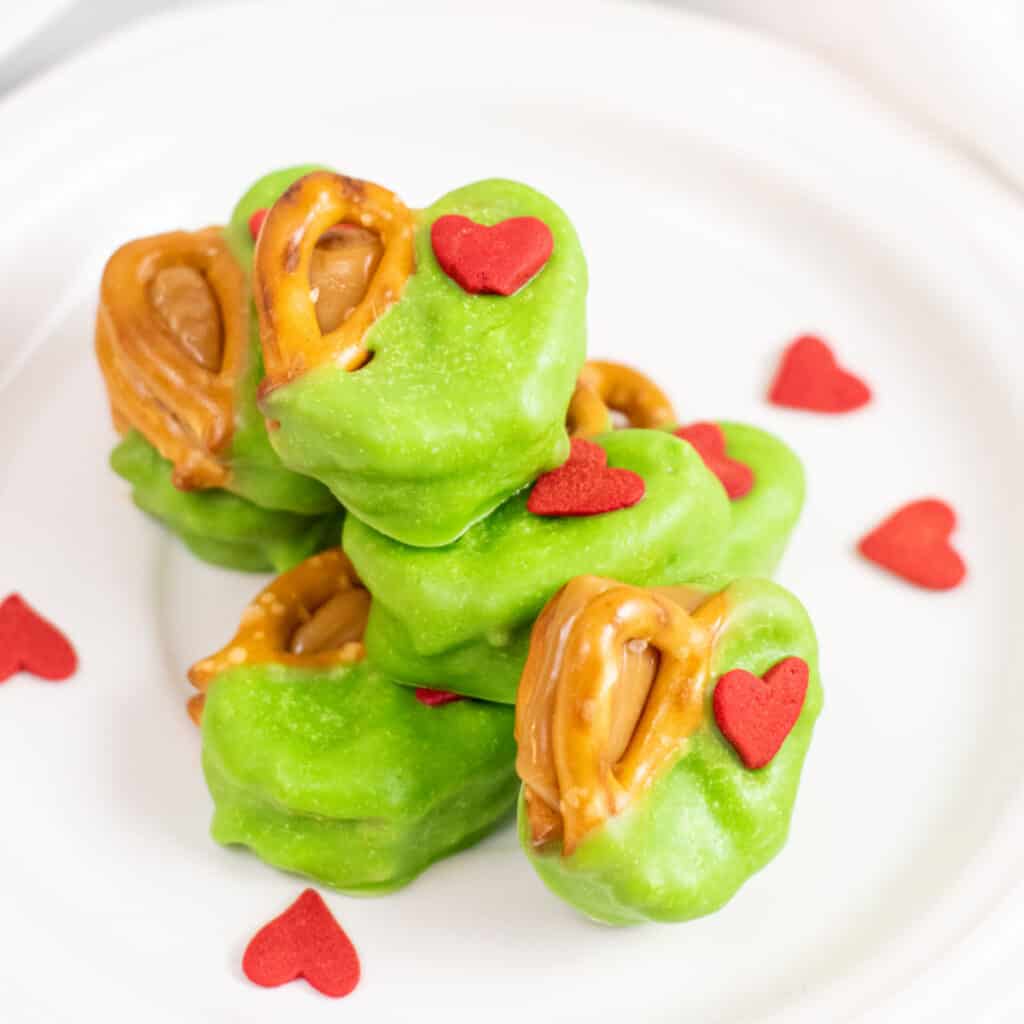 A pile of caramel filled pretzels dipped in green chocolate and decorated with  a heart shaped sprinkle