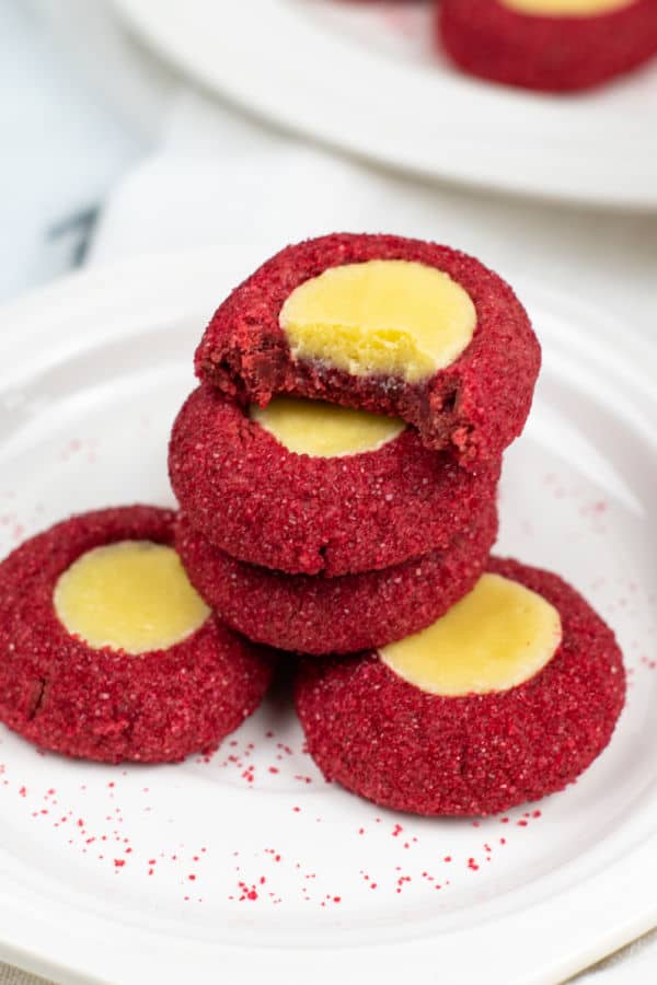 A stack of red velvet thumbprint cookies on a white plate. The top cookie has a bite taken out of it revealing the creamy texture of the center of the cookie. 