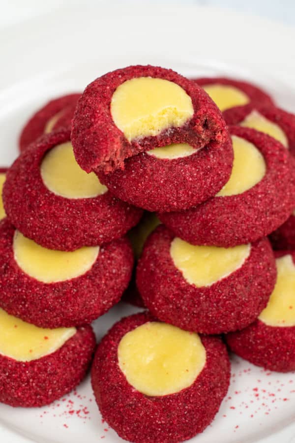 A pile of red velvet thumbprint cookies on a white plate. The top cookie has a bite taken out of it showing the creamy texture of the cookie filling. 