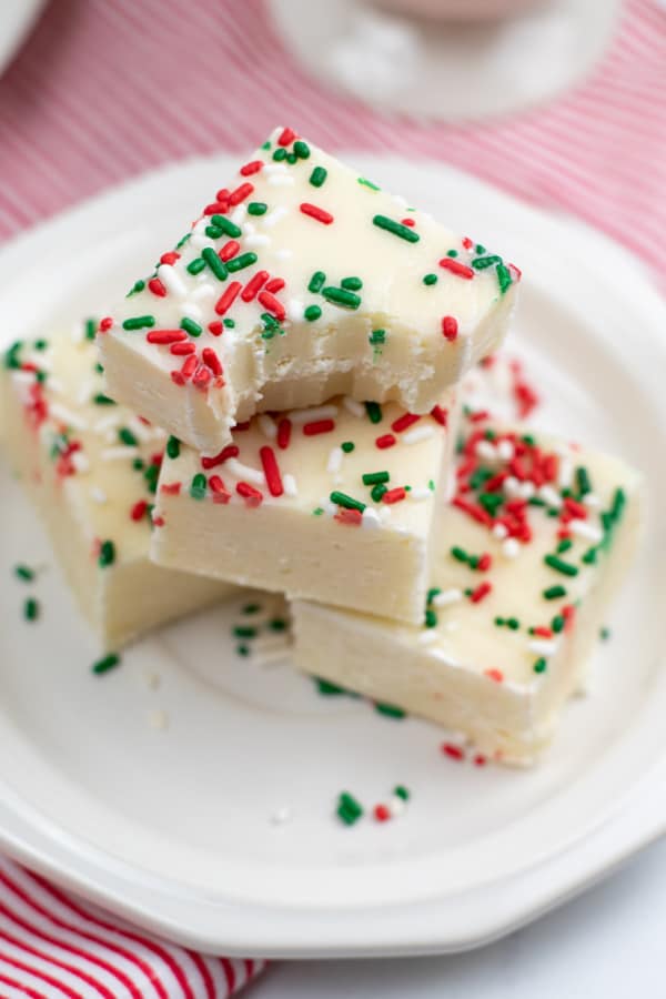 A pile of sugar cookie fudge on a white plate the top piece has a bite taken out. 