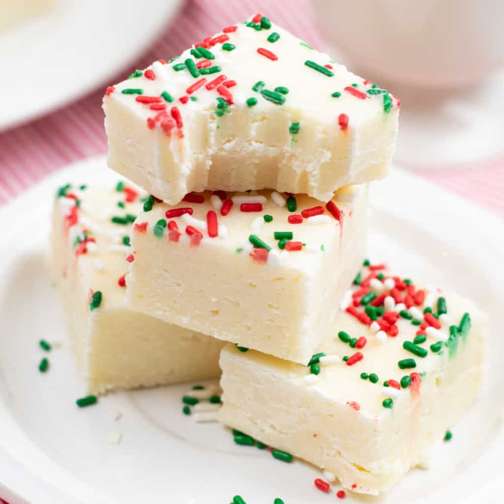 A pile of sugar cookie fudge with green, red and white sprinkles on a white plate. The top piece has a bite taken out of it. 