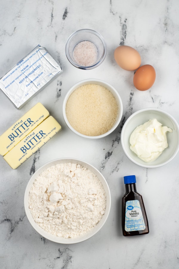 The ingredients for cheesecake thumbprint cookies on a white marble counter. Eggs, sugar, cream cheese, butter, salt, sour cream, flour and vanilla 
