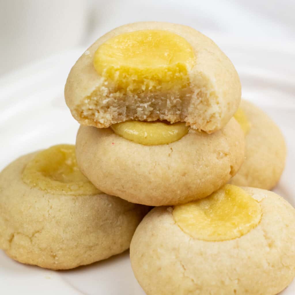 A stack of golden brown buttery cheesecake thumbprint cookies. The top cookie has a bite taken out showing the creamy cheesecake filling. 