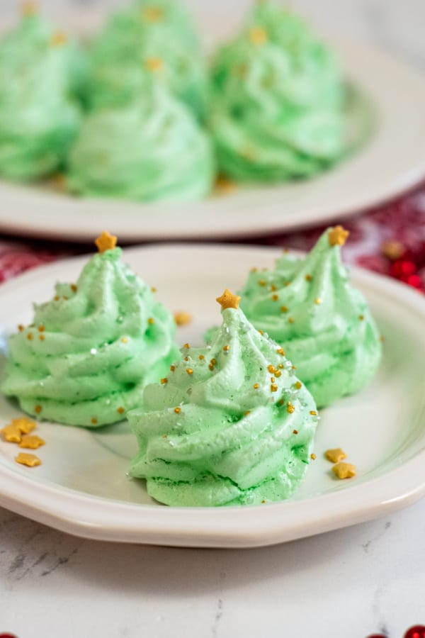 A small plate with three Christmas tree meringues . They are green and decorated with gold and silver sprinkles and topped with a gold heart. 