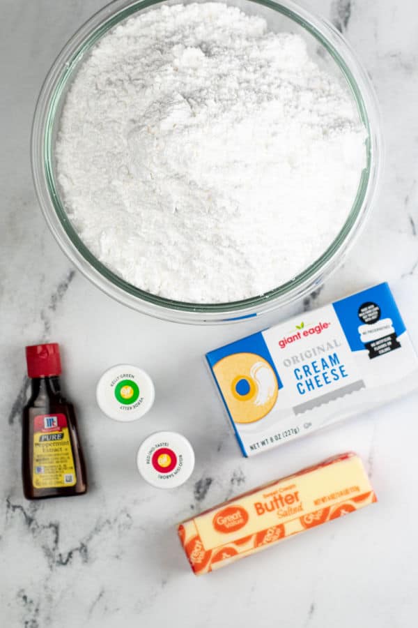 The ingredients for cream cheese mints on a white marble background. Powdered sugar, butter, cream cheese, peppermint extract and gel food coloring. 