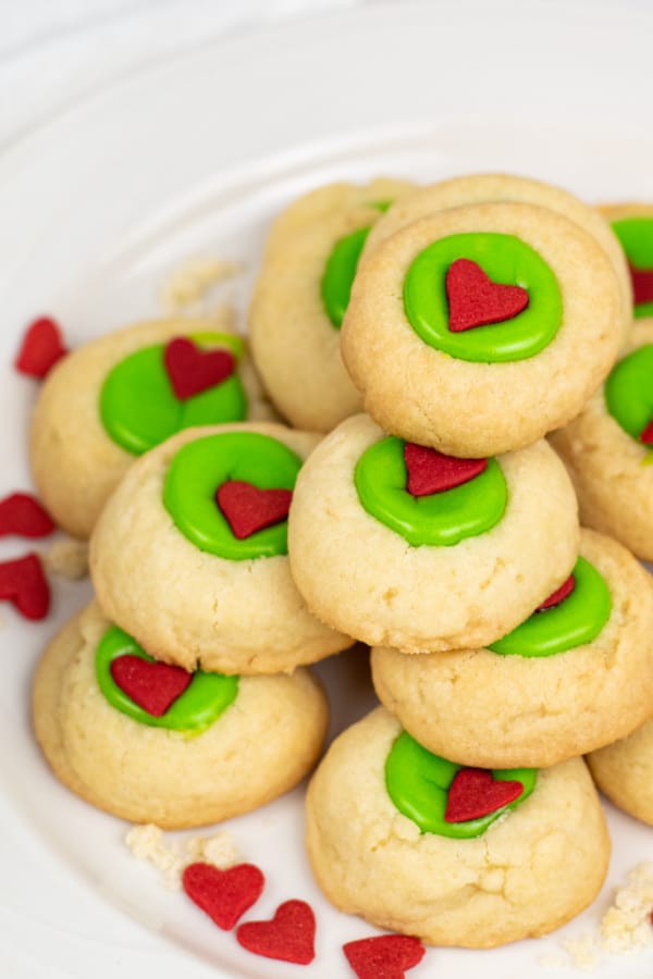 a plie of Grinch thumbprint cookies on a white plate. The cookies are golden brown with green filling and a red heart shaped sprinkle