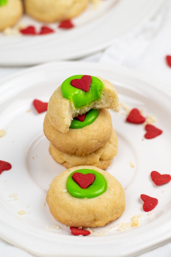 A stack of Grinch thumbprint cookies on a white plate. Golden brown cookies with green filling and red heart sprinkles. 