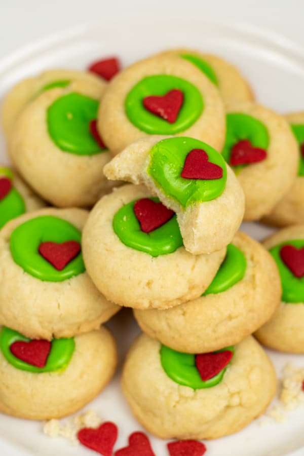 A pile of Grinch thumbprint cookies. The top cookie has a bite taken out showing the soft green center in a golden brown cookie. 