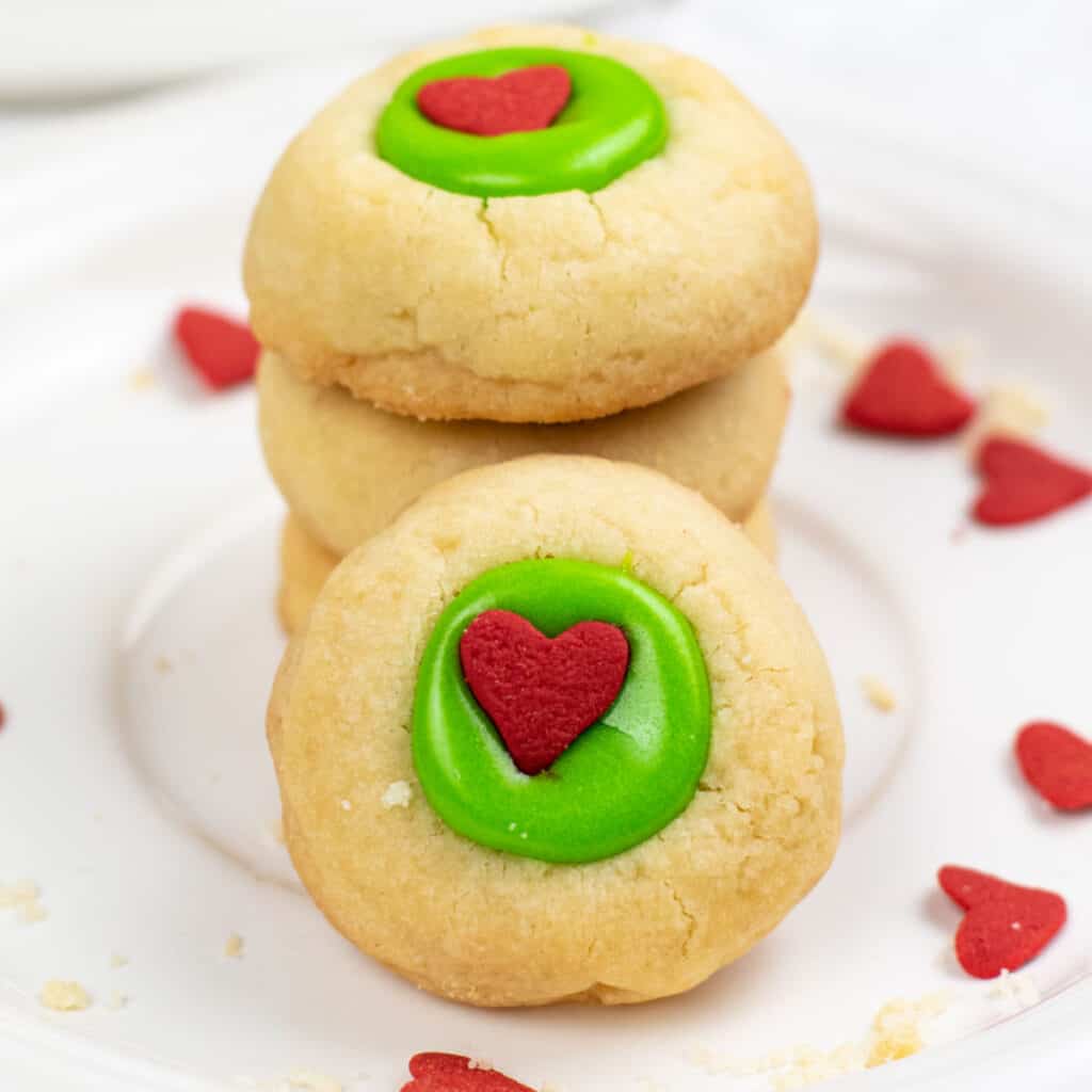 A stack of golden brown shortbread cookies filled with green frosting and topped with a red heart shaped sprinkle. 