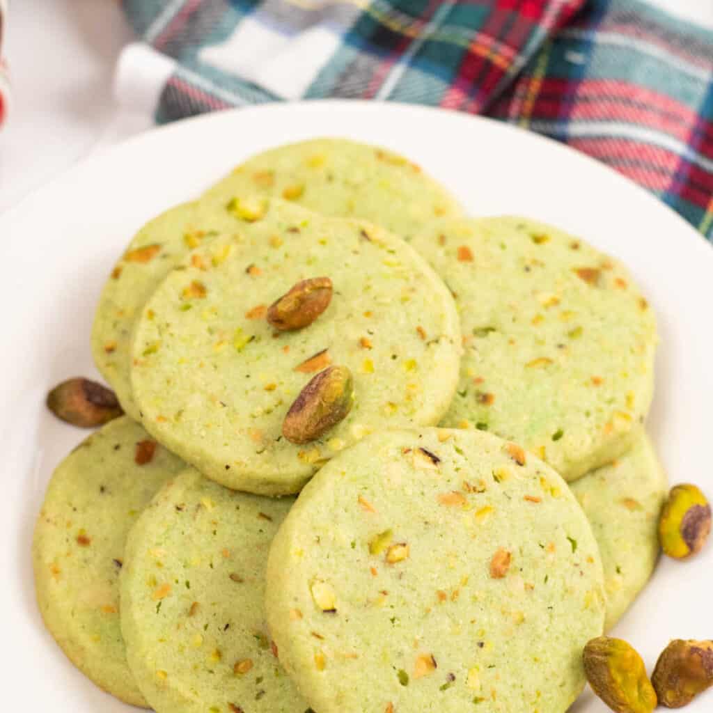 Green pistachio icebox cookies on a white plate. They are garnished with green pistachios. 