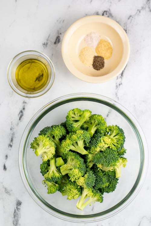An overhead view of a small glass bowl of oil, a small dish of assorted seasonings, and a large glass bowl of uncooked broccoli.