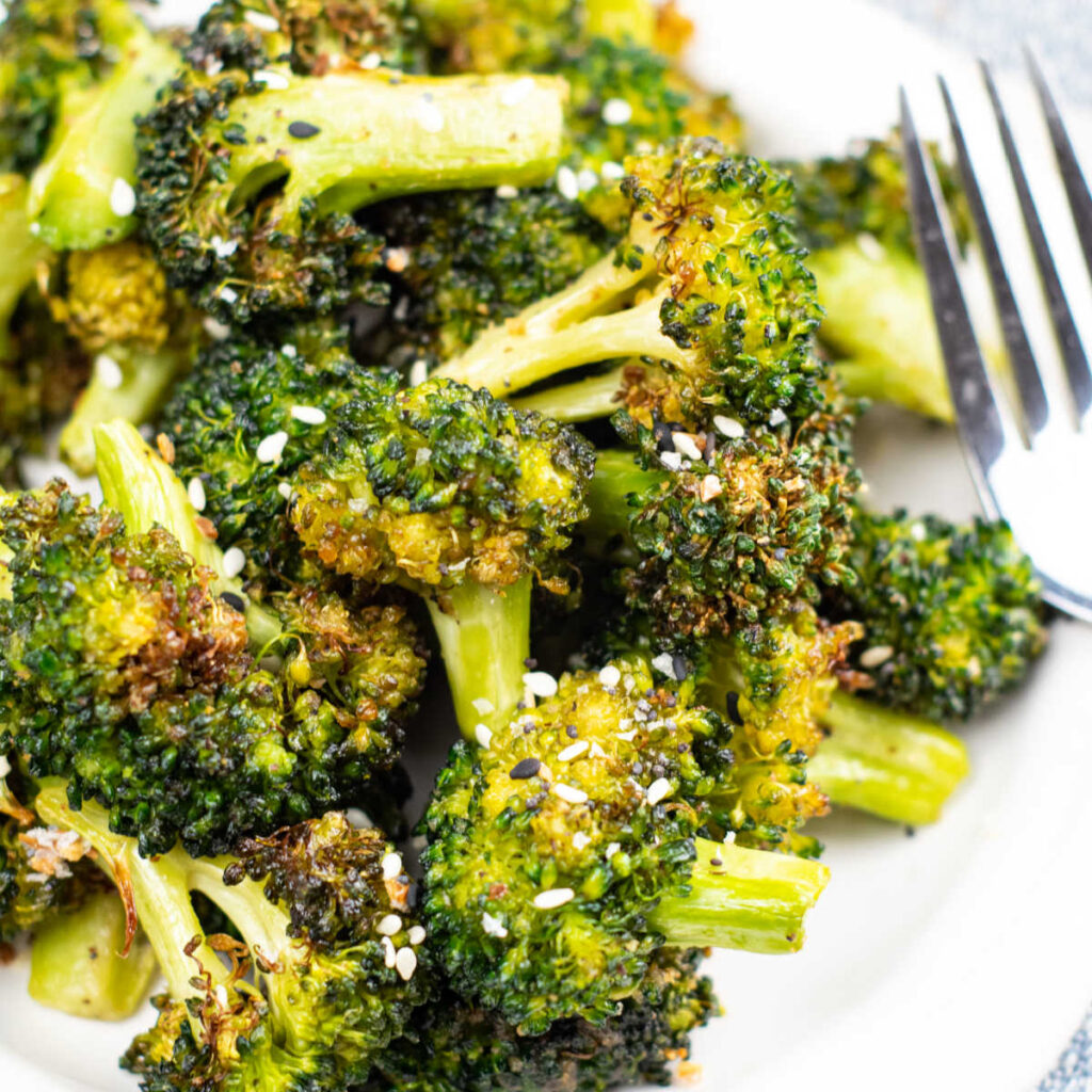 A close-up of a white plate of air fried broccoli with a fork resting on it topped with sesame seeds sitting on top of a tablecloth.