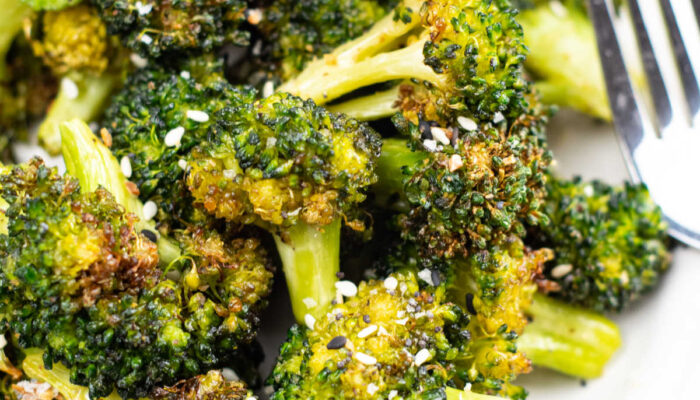 A close-up of a white plate of air fried broccoli with a fork resting on it topped with sesame seeds sitting on top of a tablecloth.