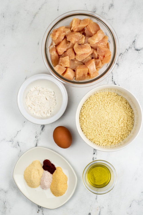 An overhead view of a large glass bowl of raw cut chicken, a small white bowl of flour, a white bowl of breadcrumbs, an egg, a small glass bowl of oil, and a dish of assorted seasonings on a marble countertop.