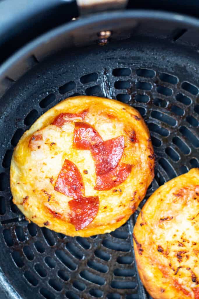 An overhead view of two mini biscuit pizzas in the basket of an air fryer, one topped with pizza sauce and cheese, the other topped with pizza sauce, cheese, and pepperoni.