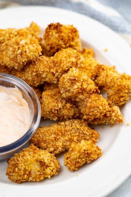 A pile of cooked popcorn chicken and a small glass bowl of creamy dipping sauce sitting on a white plate on top of a tablecloth,