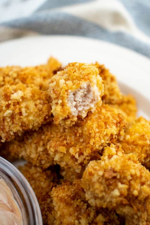 A close-up of a pile of cooked popcorn chicken, one piece with a bite out of it, sitting on a white plate on top of a tablecloth.