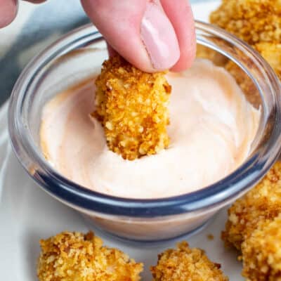 A close-up of a hand dipping a cooked piece of popcorn chicken into a small glass bowl of creamy dipping sauce on top of a tablecloth.