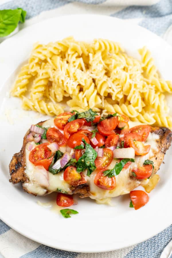 A plate with  cooked chicken breast with tomatoes, basil and mozzarella cheese on it and a pile of rotini pasta. 
