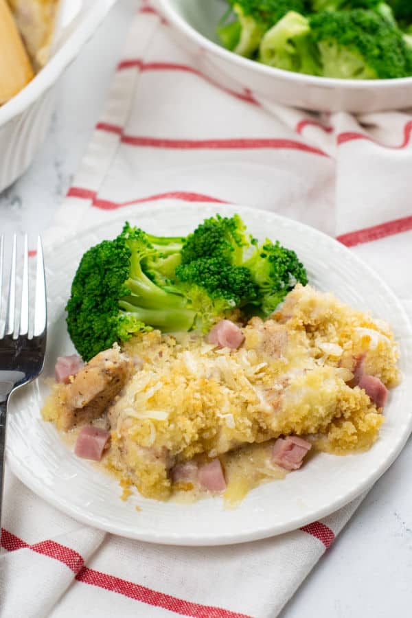 A plate of cooked chicken cordon bleu and broccoli with a fork resting on top sitting on a dish towel on a marble countertop.