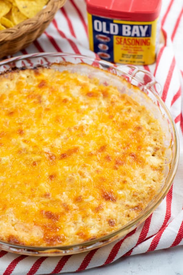 A clear glass pie pan with cheesy crab dip in it, in the background is a container of old bay and a basket of tortilla chips. 