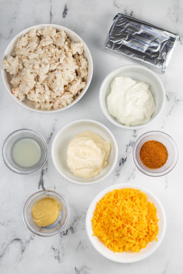 The ingredients for Maryland crab dip on a white marble counter top. Lump crab meat, cream cheese, sour cream, mayonnaise, lemon juice, old bay seasoning, dijon mustard, shredded cheddar cheese. 