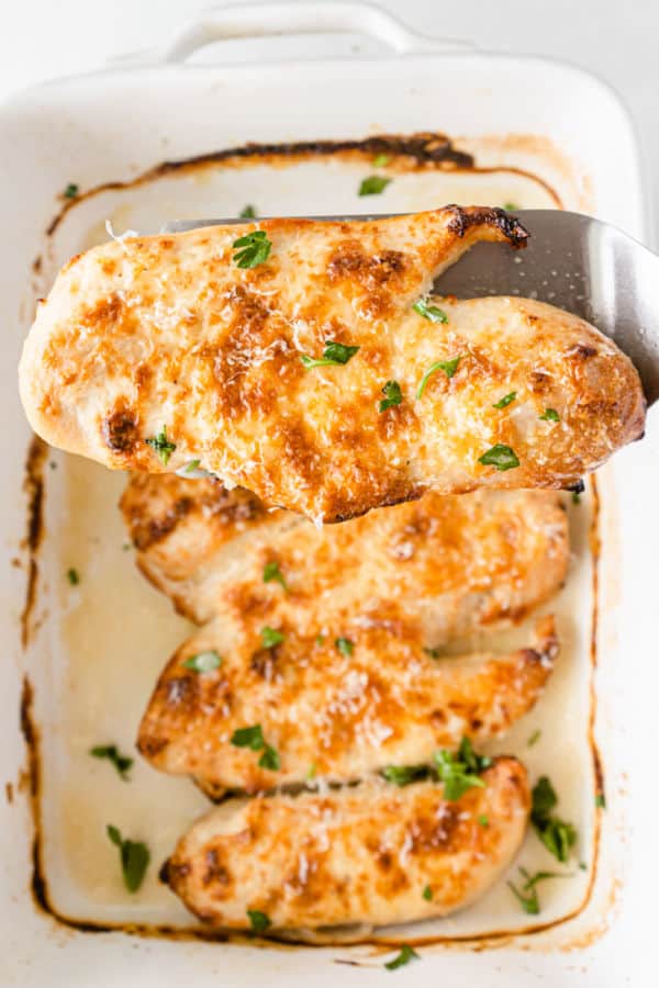 A white casserole dish full of golden brown parmesan mayo chicken garnished with chopped parsley. 