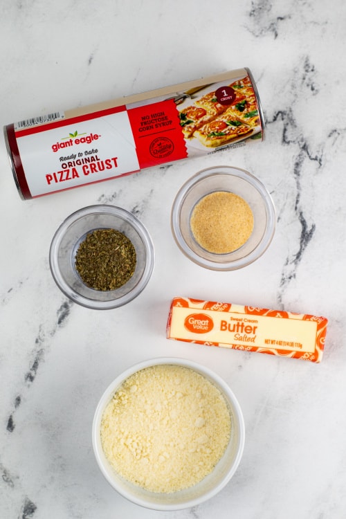 An overhead view of a can of raw pizza dough, a small glass bowl of Italian seasoning, a small glass bowl of garlic powder, a stick of butter, and a large white bowl of Parmesan cheese on a marble countertop.