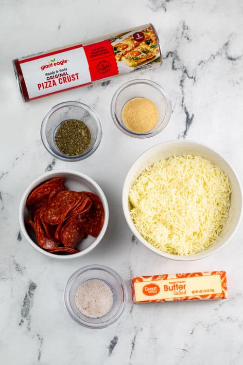 An overhead view of several bowls of ingredients for air fryer pepperoni rolls, along with a stick of salted butter and a tin of uncooked pizza crust on a marble countertop.