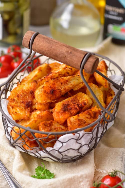 A metal basket of air fryer potato wedges on top of a beige linen cloth, with various other ingredients in the background.