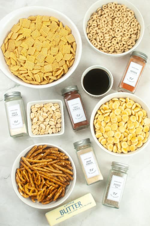 An overhead view of several white bowls of ingredients for air fryer chex mix, along with several glass containers of seasoning, and a stick of salted butter on a marble countertop.