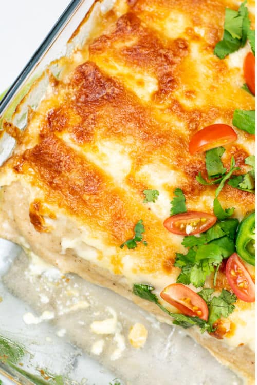 An overhead view of a large glass baking tray of creamy chicken enchiladas garnished with jalapeños, cherry tomatoes, and cilantro on a marble countertop.