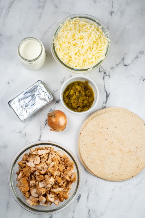 An overhead view of a glass of heavy cream, a large glass bowl of shredded cheese, several tortillas, a large glass bowl of chicken, and several other ingredients on a marble countertop.