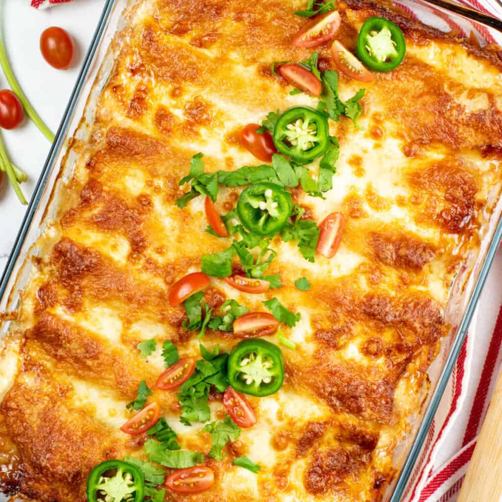 An overhead view of a large glass baking tray of creamy chicken enchiladas garnished with jalapeños, cherry tomatoes, and cilantro on a marble countertop.