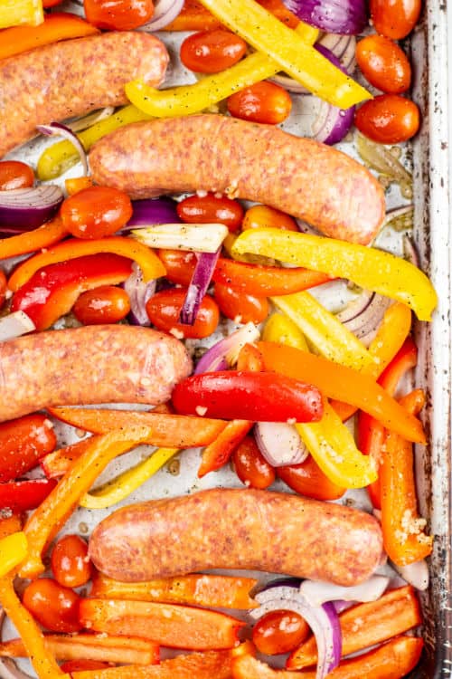 An overhead view of a sheet pan of uncooked Italian sausage and various mixed vegetables.
