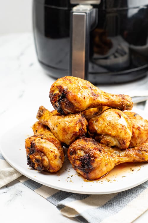 A white plate of air fried chicken legs on a marble countertop in front of an air fryer.
