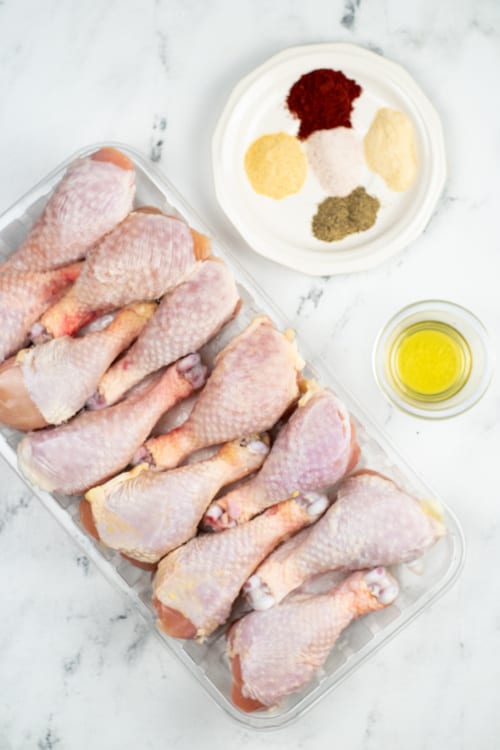 An overhead view of a container of raw chicken legs, a plate of seasoning, and a small glass bowl of oil on a marble countertop.