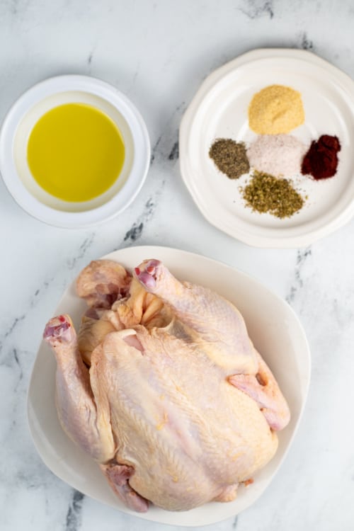 The ingredients for air fryer rotisserie chicken on a white marble counter top. A whole chicken, olive oil, and spices.