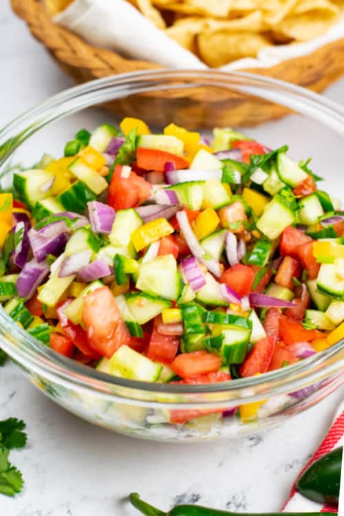 A large glass bowl of cucumber pico de gallo on a marble countertop.