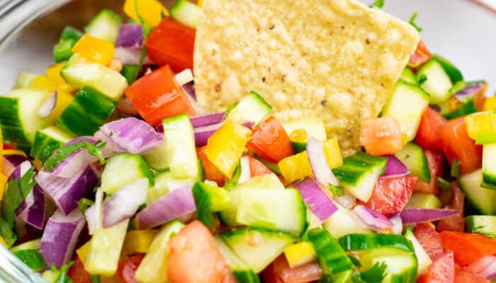 A close-up of a tortilla chip dipped into a large glass bowl of cucumber pico de gallo.
