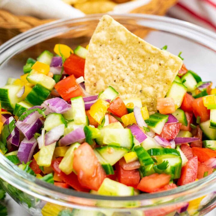 A close-up of a tortilla chip dipped into a large glass bowl of cucumber pico de gallo.